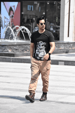 Man wearing a black t-shirt with a graphic design, beige pants, and sunglasses standing in front of a fountain.