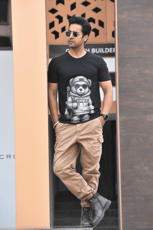 Man wearing a black t-shirt with a graphic design, beige pants, and sunglasses standing against a textured wall.