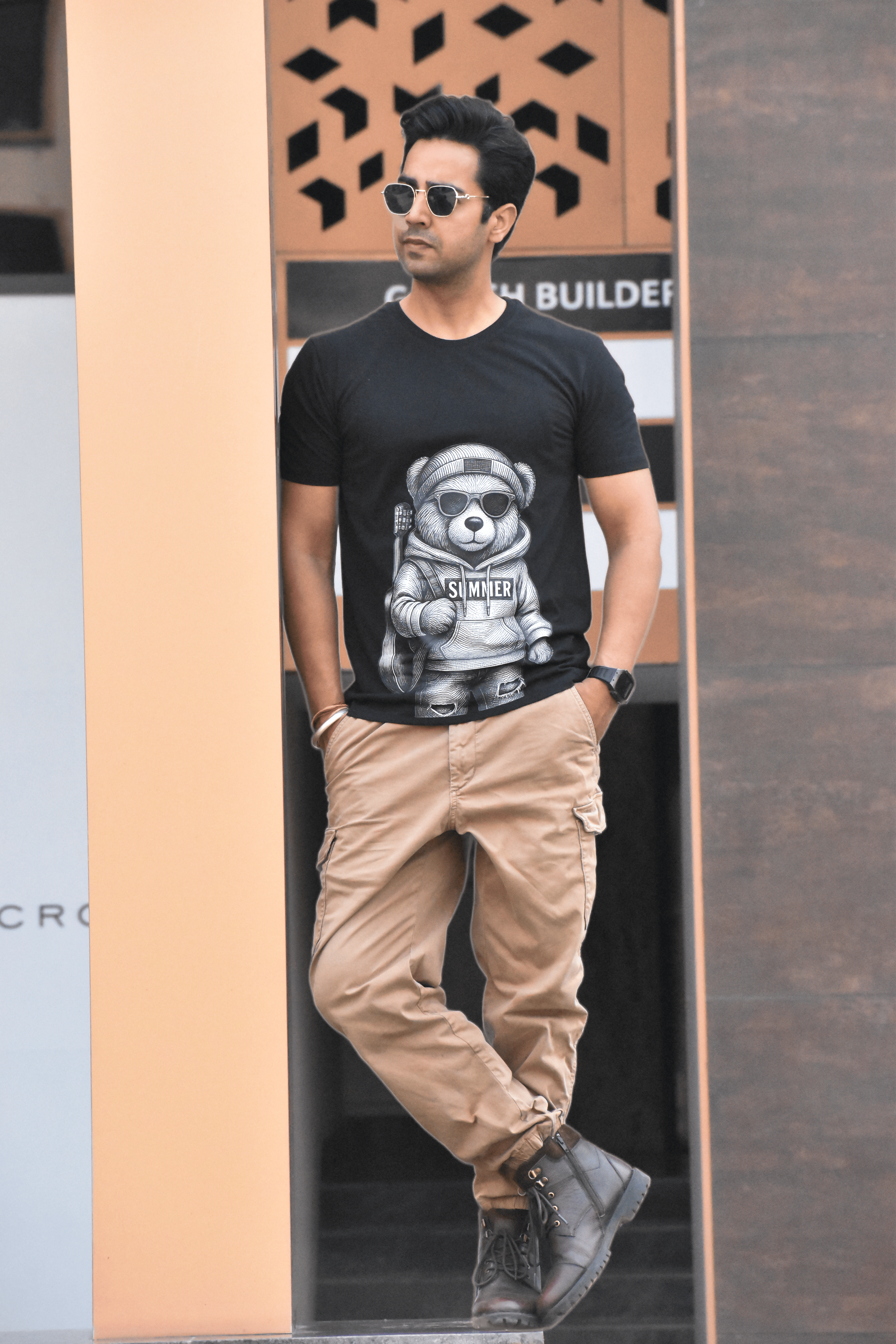 Man wearing a black t-shirt with a graphic design, beige pants, and sunglasses standing against a textured wall.